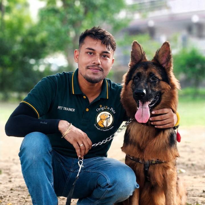 Origizn Pets trainer crouched with arm around a long-haired German Shepherd outdoors in a green park setting